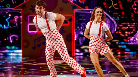 George and Alexis on the dancefloor, dressed in love-heart jumpsuits and matching red shoes. They both have one leg out to the side as they dance. Brightly-coloured shapes are projected onto the dancefloor and walls.