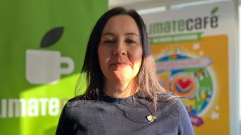BBC Jess Pepper, a dark-haired woman in a black sweater and a Climate Café pin badge, stands in front of a the café signage which depicts a coffee mug and a colourful 10-year anniversary graphic.