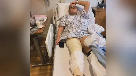 Hudgell Solicitors A man in a hospital robe lying in bed with a bandage covering his right leg. He wears glasses and has a mobile phone by his side. 