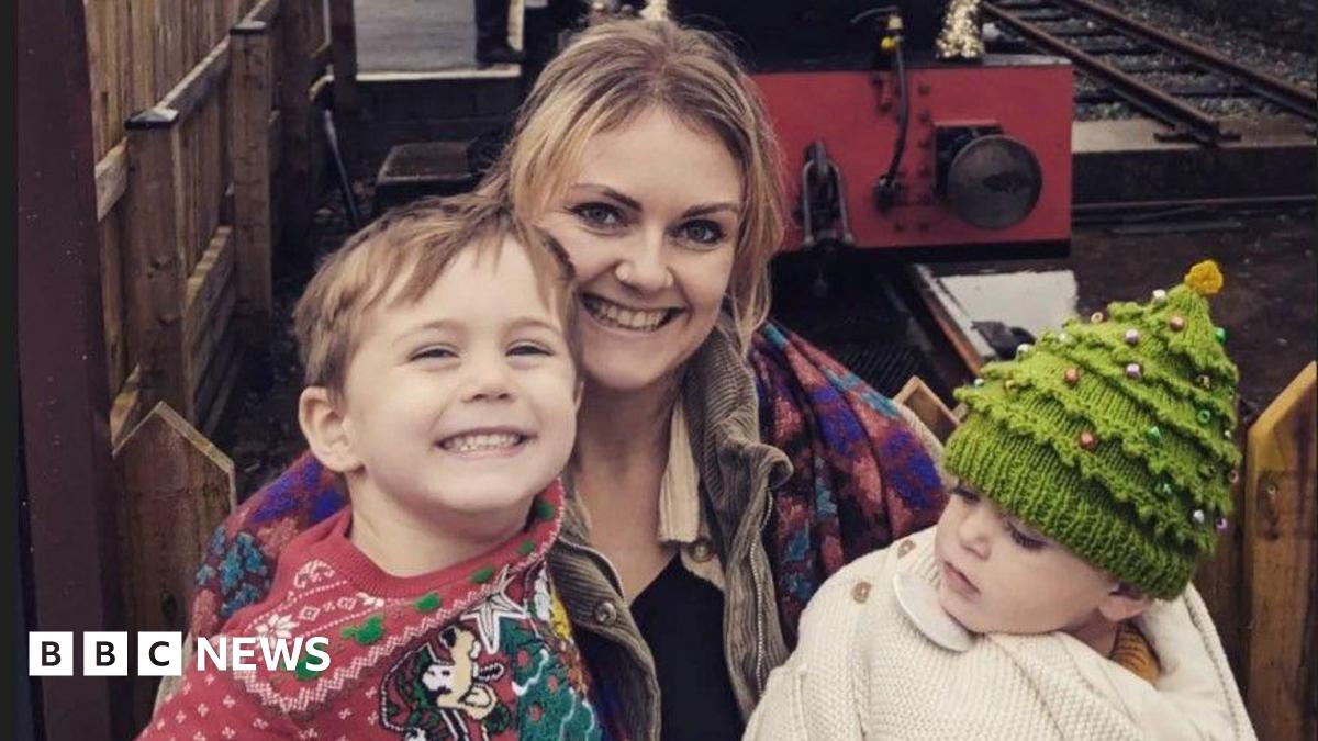 A woman with long blonde hair is smiling while holding a young boy and a baby boy. They are sitting by a mini train. The older boy is smiling and wearing a red Disney-themed Christmas jumper and the baby has a Christmas tree-style woollen hat.