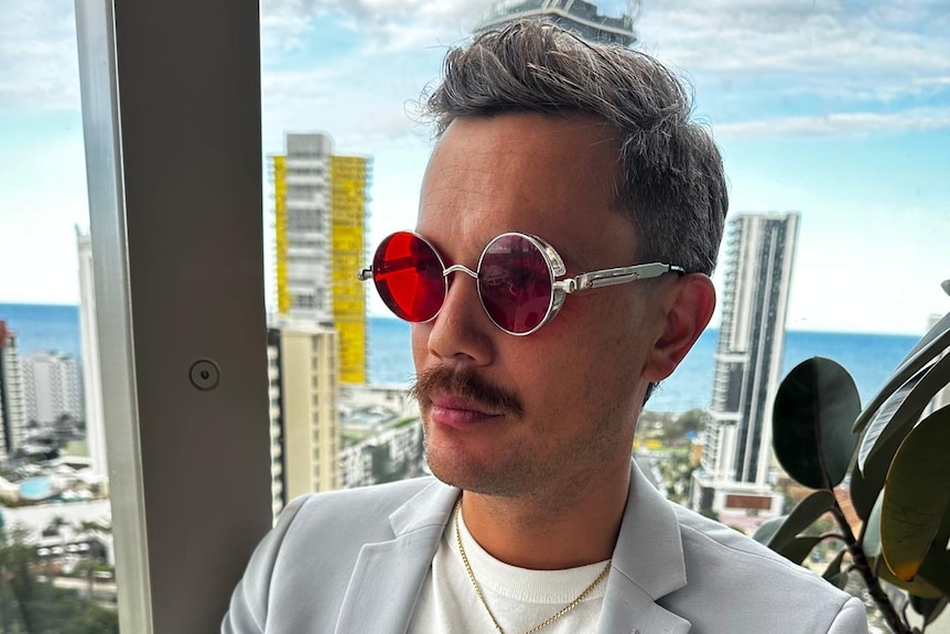 A man in a white tee and blue blazer, moustache and silver sunglasses sitting in a restaurant.