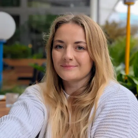 A selfie of Dr Tayla Hammond, animal welfare research and policy consultant. She is a blonde-haired young woman sitting at an outdoor table and wearing a white ribbed sweater.