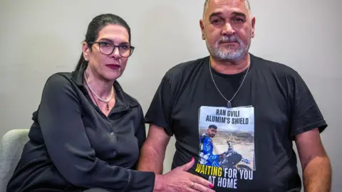 Talik, who has black hair in a ponytail and wears black-rimmed glasses, poses for a photo with her husband Itzik, who has grey short hair and a beard and wears a silver chain. They both have neutral expressions.