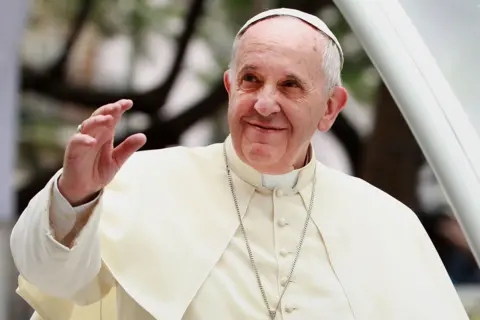 Getty Images Pope Francis is wearing a cream-coloured robe with a matching skullcap and a long silver chain with a cross pendant. One hand is raised in a gesture, while the other is partially visible. The background shows blurred greenery and architectural elements, suggesting an outdoor setting at a formal or ceremonial event.