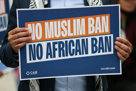 AFP via Getty Images A man holds a navy blue and orange sign with white lettering that reads 'No Muslim Ban, No African ban'. 