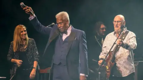 Getty Images Eddie Floyd, middle, raises a microphone in his right hand, with Cropper playing guitar to his left, and Sheryl Crow to his right