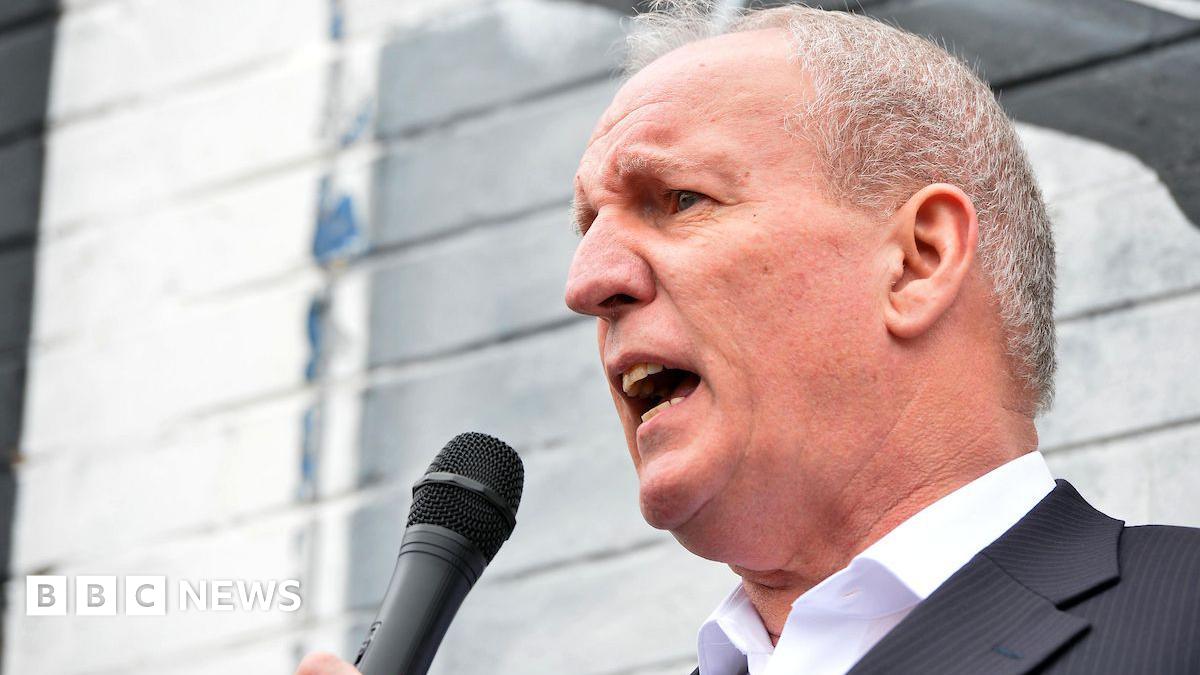 Bobby Storey is speaking into a mic. He's outside. A painted wall is behind him. He's wearing a black suit with a white shirt.