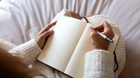 Getty Images High angle view of unrecognizable young woman wearing a robe sitting on bed writing on journal in cozy bedroom