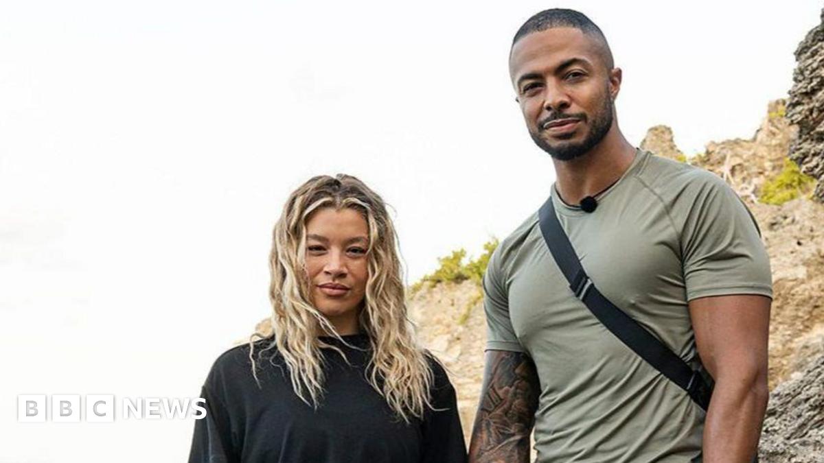 Tyler West and Molly Rainford hold hands and smile stood on rocks by the shore of the sea.
