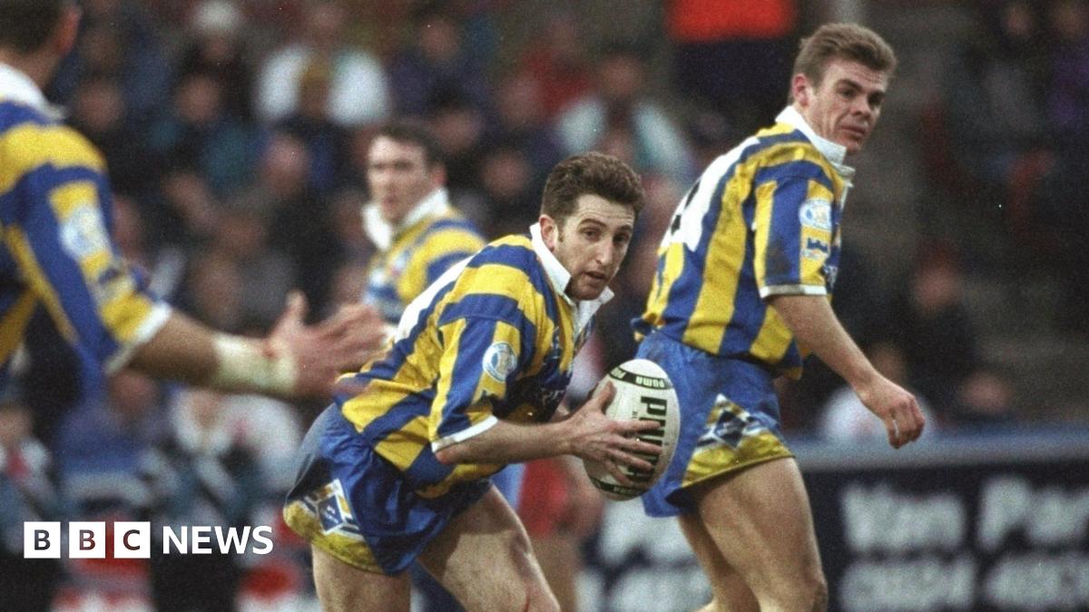 A woman with long light brown hair, dark brown eyes and fringe is smiling into the camera. She has a green, cream and black chequered shirt on. The background is white with nothing else in frame. She has a long blue decoration on her right ear