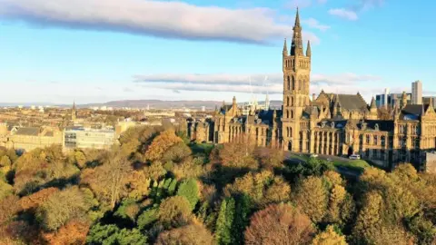 Getty Images Glasgow University, a gothic building with a tall tower, stands on a hill above trees at Kelvingrove Park overlooking buildings beyond the campus, with hills in the distance.