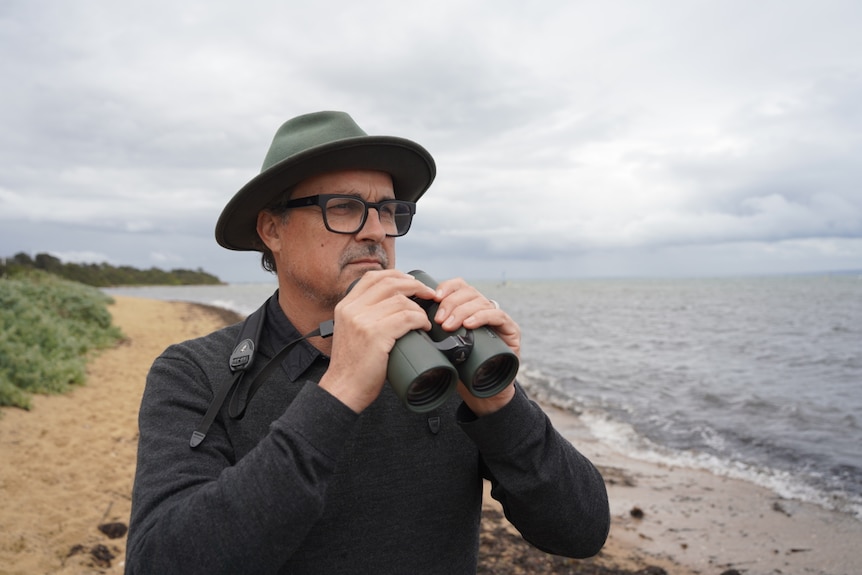 Bird Enthusiast Sean Dooley holds a pair of binoculars at the beach, looking ahead.