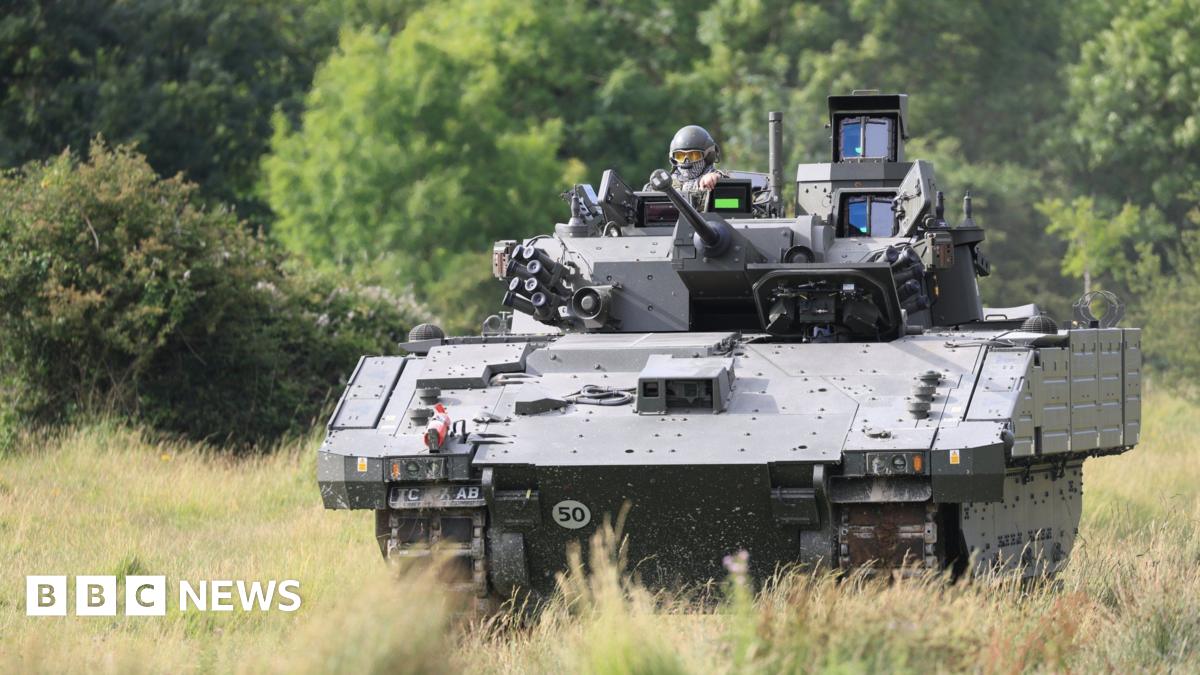An AJAX armoured fighting vehicle, a large green tank, is seen being driven through a field.