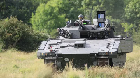 MOD An AJAX armoured fighting vehicle, a large green tank, is seen being driven through a field.