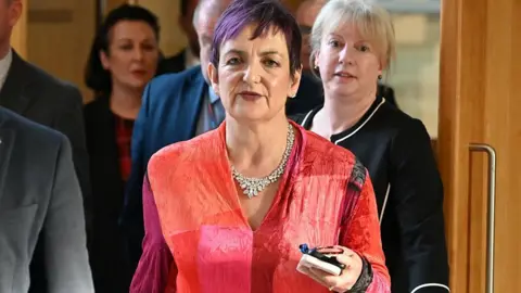Getty Images Angela Constance, who has short purple-dyed hair and is wearing a red shirt, is walking through the Holyrood building, flanked by Finance Minister Shona Robison and some men in grey suits.