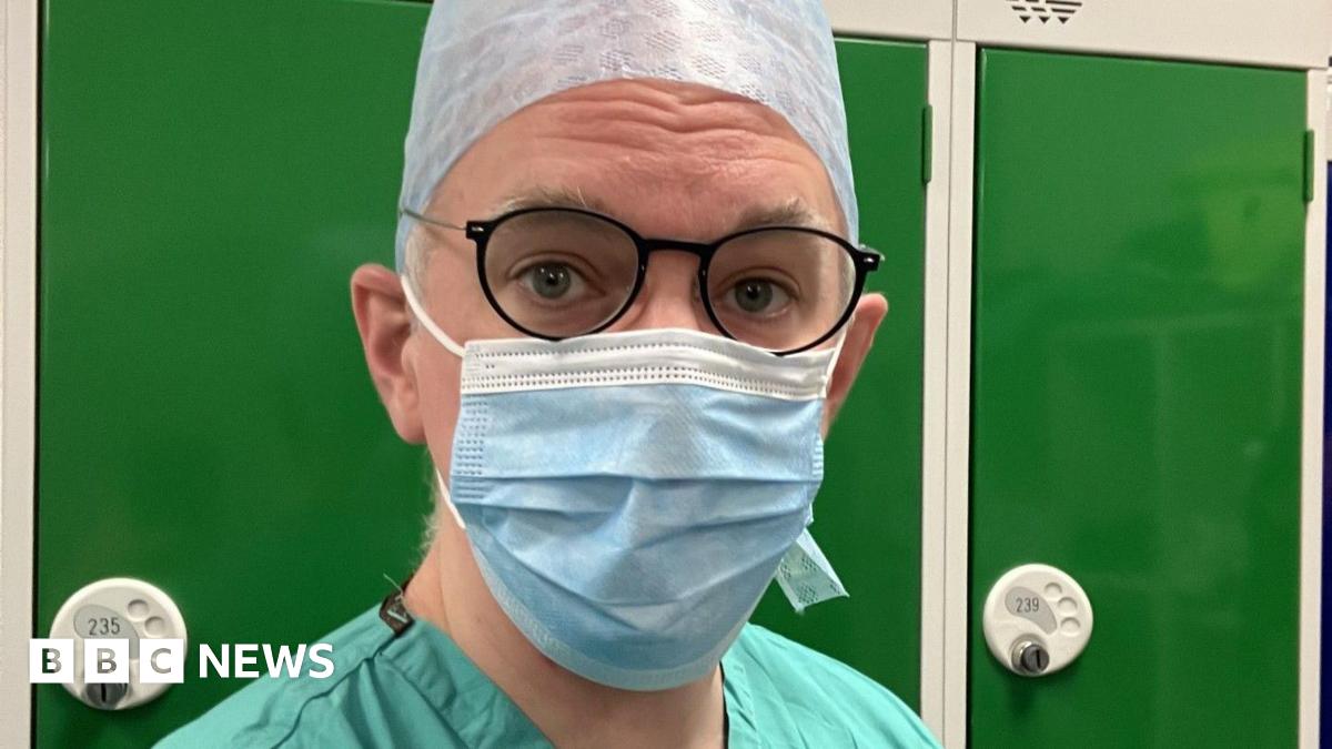 Head and shoulder selfie of correspondent James Gallagher, in green surgical scrubs, a blue facemask and a blue surgical hat. He is wearing dark blue glasses with metal arms. He is standing in front of a row of dark green lockers