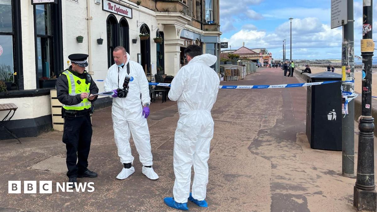 Two men in forensic suits, one with a camera hanging from his neck, stand on Portobello beach promenade talking to a police officer, who is in full uniform and also wearing a hi-vis jacket. There is the sandy beach on one side of them and a large building on the other with the name The Esplanade on it.