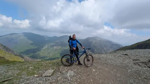 Phil James A man in blue sportswear is standing next to a bike on Mount Snowdon. There are hills in the background and he is wearing a rucksack and blue long sleeved top.