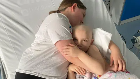 Family Ellie and Emilia are hugging as they are reunited in hospital after a long time apart. They are lying on Emilia's hospital bed with their arms around each other. Emilia is looking happy to be back with her big sister.