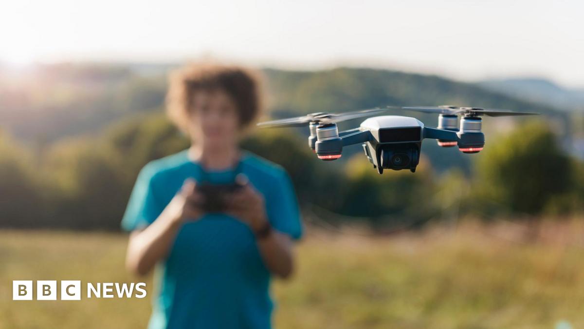 A man flies a drone in a field, surrounded by hills and greenery on a sunny evening.