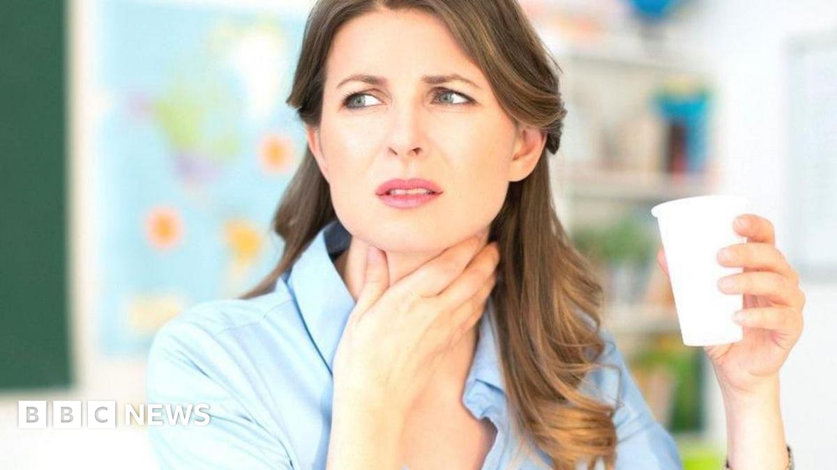 A woman with long brown hair and wearing a blue blouse clutches her throat with her hand, looking pained, while holding a white cup of something to drink