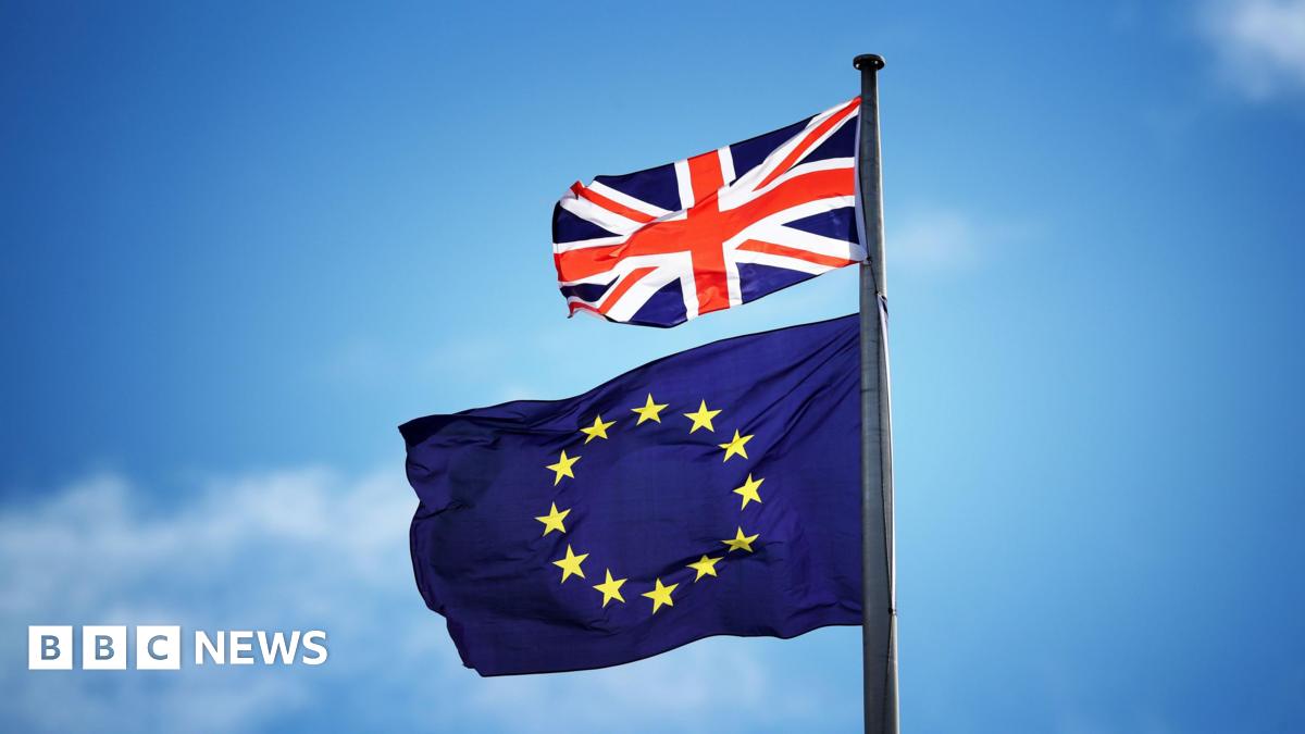 A small red, white and blue British Union Jack blows in the breeze above a a European Union flag, which consists of a ring of yellow stars against a block blue background. The sky in the background is clear.