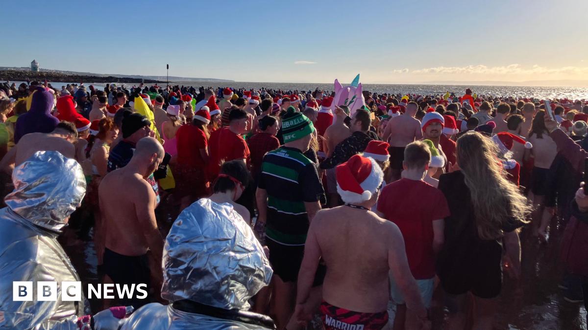 A large crowd from the behind getting ready to enter the water for a sea dip. Many are wearing Santa hats and festive costumes. The sun is shining.