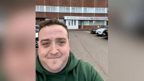 Stephen Halsey Stephen Halsey with short brown hair wearing a green hoodie standing in front of the children's outpatients department at Arrowe Park Hospital. He is smiling.