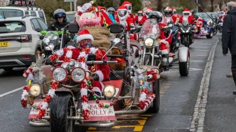 Del Hickey A group of Santas, all wearing the classic red and white suits, ride on decorated motorbikes down a street as onlookers watch. 