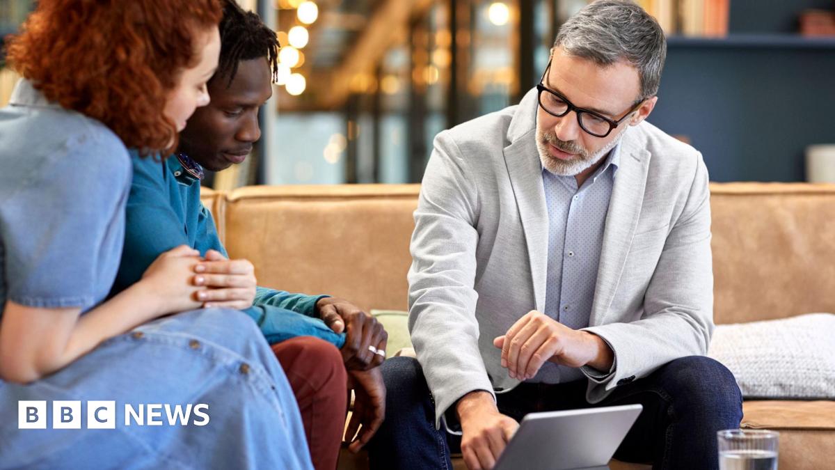 Man and woman sit on a sofa where they are shown something on a tablet computer by a man in a blazer and shirt.