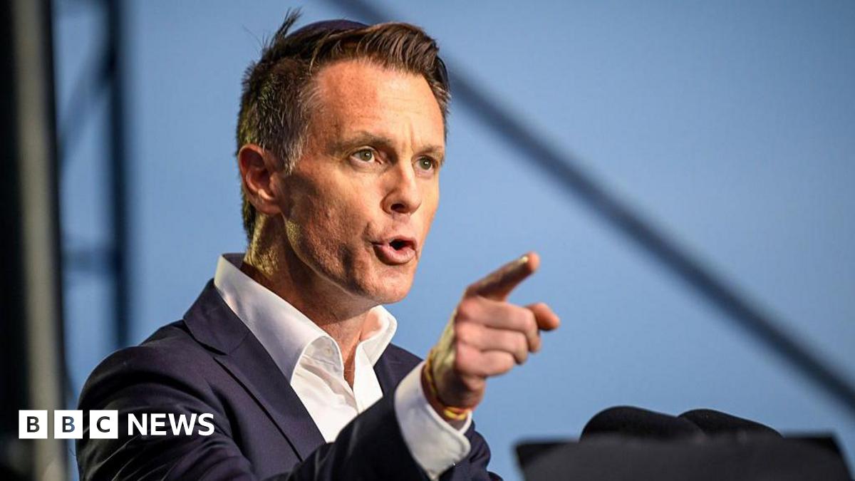 A man in a dark suit speaking and gesturing with his hand, pointing his index finger