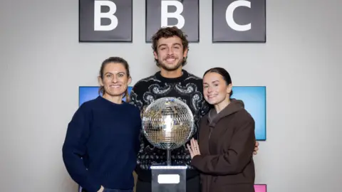 Guy Levy/PA Wire Karen, George and Amber are stood behind the glitterball trophy with the BBC logo on the wall behind them.
