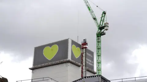 PA Media Grenfell Tower partially covered in grey cladding with green heart symbols, as a crane stands beside the building during deconstruction work.