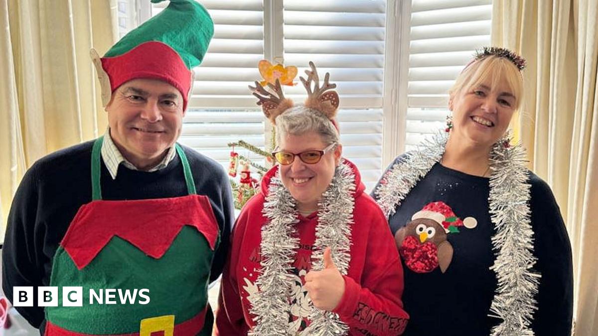 Left to right: Mike, Clare and Kim. Mike is wearing an elves apron and hat. Clare has a red Christmas jumper with silver tinsel round her neck and reindeer ears. Kim with blonde hair wears a navy blue Christmas jumper and silver tinsel around her neck. They are all standing in front of a Christmas tree and smiling.