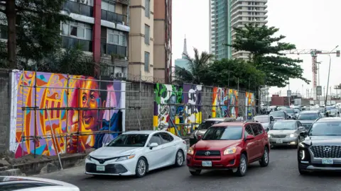Toyin Adedokun / AFP via Getty Images Several brightly coloured murals are seen on a wall next to a busy road full of cars. A crane and a tower bloc can be seen in the distance. Behind the wall are trees and a block of flats.