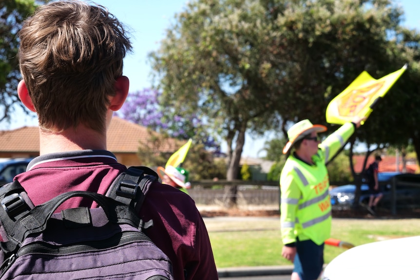 A male high school students waits to cross a road while a traffic warden in high-vis waves a flag.