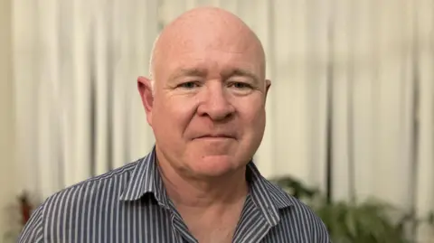 BBC/Lucy Parry A head-and-shoulders image showing Steve Foster looking directly at the camera. He is semi-bald, with closely cropped white hair. He wears a striped blue and white shirt. He is standing in front of a beige window blind and there are pot-plants on a shelf behind him.