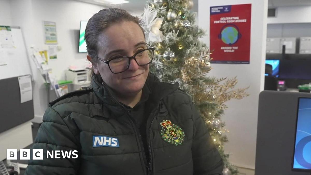 An London ambulance worker standing in a call centre. She is wearing a green jacket and there is a Christmas tree behind her.