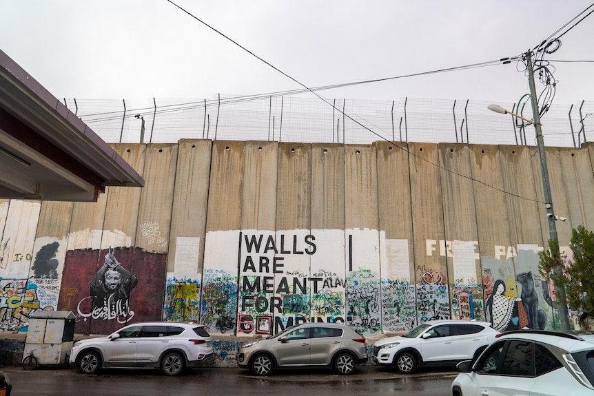 A large long wall, with wire on top, has graffiti on it and cars parked next to it. 