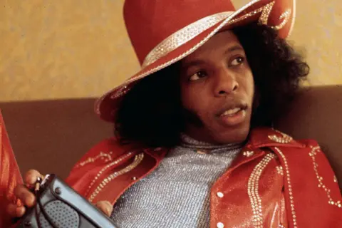 Getty Images Sly Stone is dressed in a shiny silver top paired with a vibrant red leather jacket and a matching wide-brimmed hat. Both the jacket and hat are decorated with sparkling embellishments arranged in patterns. Sly is holding a gray rectangular object with a speaker grille and buttons, possibly a portable radio or electronic device. The background features a textured beige wall, suggesting an indoor setting.