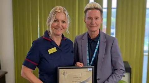 RJAH A nurse receiving an award, stood next to a man in a suit. they are both holding a frame with a certificate inside 