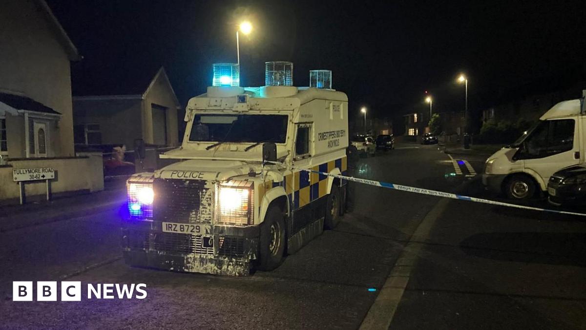 A police vehicle sits in the middle of a road. It is dark. The large vehicle is white. A police cordon tape is across its bonnet. A road sign reading Tullymore Road is to its left