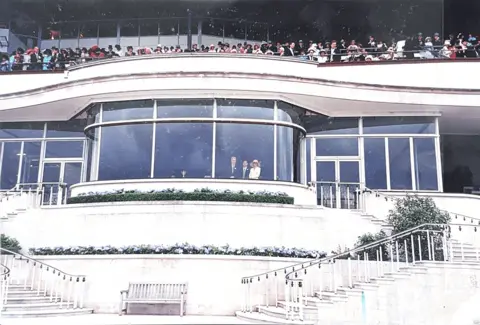 US Department of Justice A wide shot of a large white building, with glass windows, with Epstein, Andrew and Maxwell standing at a window. Above them are crowds of people.
