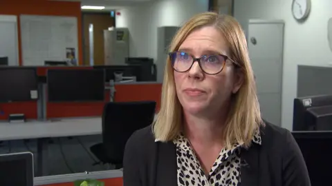 BBC Detective Inspector Cheryl Lewendon is sitting in a police station office with a row of computers behind her