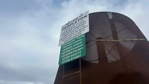 A white sign reads "Welcome to Peopleton. A beautiful village populated by a caring Godfearing community a great place to live probably most definately the nicest village in Worcestershire (sic)." A green sign beneath it reads "Stonearrow Farm" along with some contact numbers. 