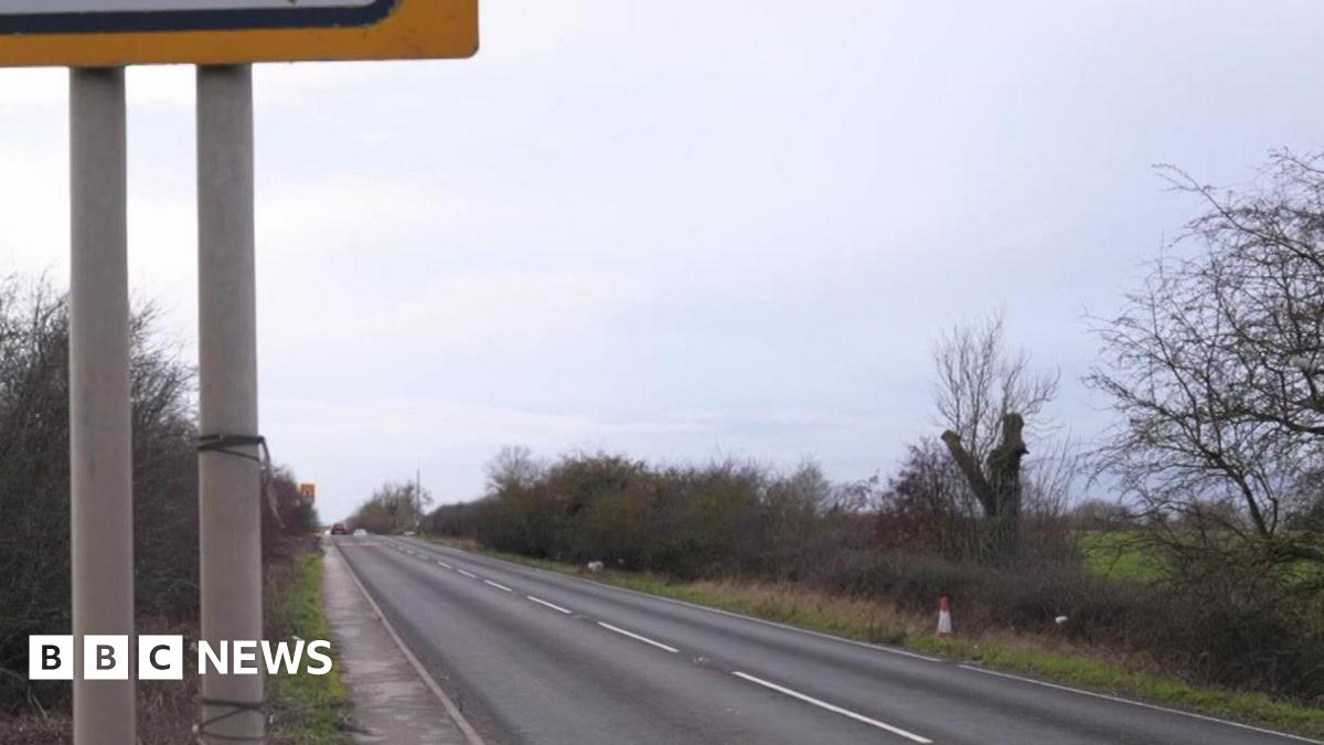 A rural road bordered by grassy verges and leafless trees. A black car is driving away in the distance. On the right side of the image, a white street sign reads “TODWICK ROAD S26” near a junction with traffic islands and bollards.