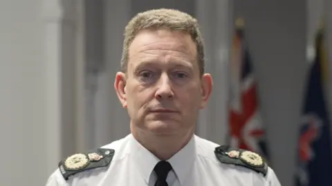 Jamie Niblock/BBC Ben-Julian Harrington is wearing a white shirt and black tie with black and gold epaulettes on his shoulders. He has short brown hair and looks at the camera with a serious expression. Behind him are a Union Jack flag and a navy Essex Police flag.
