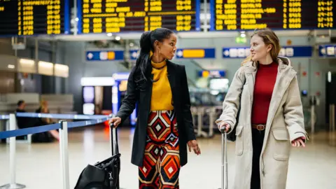 Getty Images Two ladies carry their suitcases through an airport with a full departure board behind them