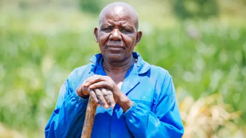 Ali Ngwane / BBC A portrait of Frederick Bwalya, 73, a farmer in Twalima village, standing against a sunny and grassy backdrop. He looks pensively into the camera, resting his hand on a wooden stick and is wearing a bright blue shirt jacket. 