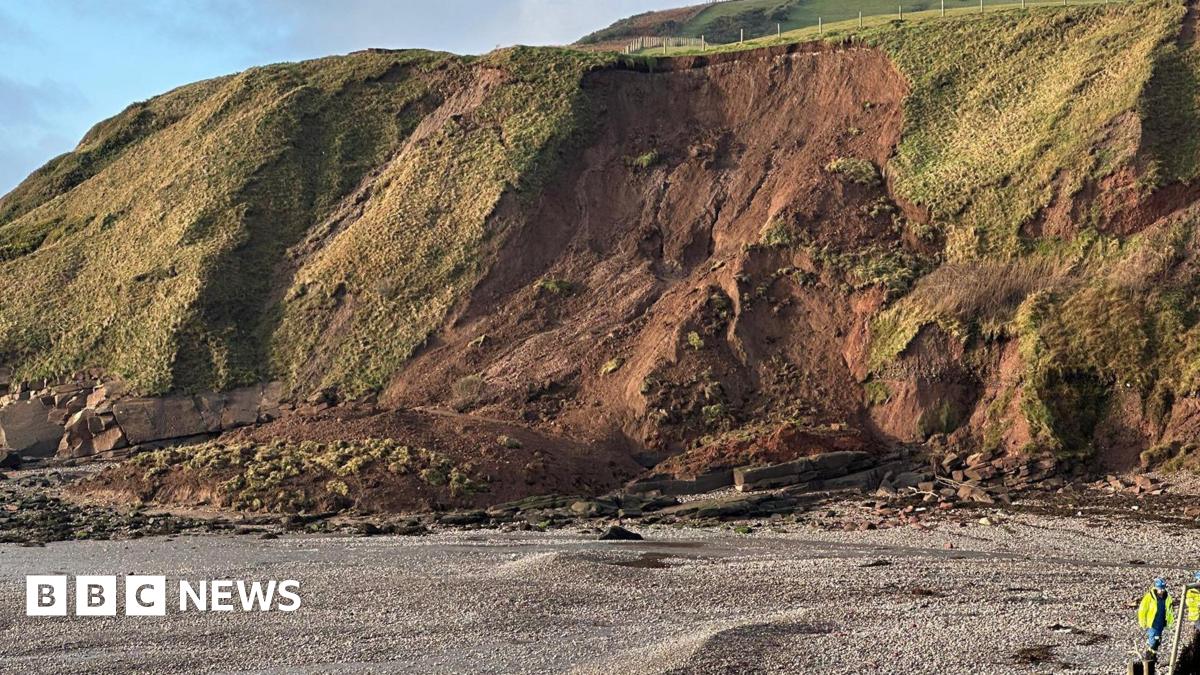 A large landslip on a beach-front cliff. Mud is piled at the bottom of the grassy cliff face. Two people in high-vis jackets are standing on the stone beach.